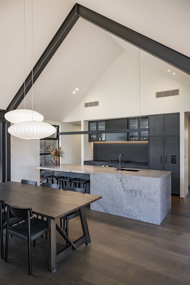 A sophisticated open-plan kitchen and dining area with a dramatic vaulted ceiling and exposed dark structural trim. The kitchen features a large grey stone island with waterfall sides, dark integrated cabinetry, a dark tiled backsplash, and concealed warm under-cabinet lighting. A dark timber dining table with black chairs sits in the foreground beneath sculptural white pendant lights. The space has dark timber flooring, pale walls, black-framed glazing, and a refined contemporary material palette.