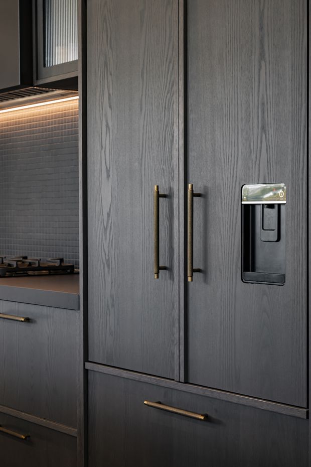 Close-up view of a sophisticated dark kitchen featuring tall grey wood-grain cabinetry, integrated refrigerator doors with brass bar handles, a built-in water dispenser, a gas cooktop, and a dark small-format tiled splashback. Warm under-cabinet lighting adds contrast to the moody, refined material palette.