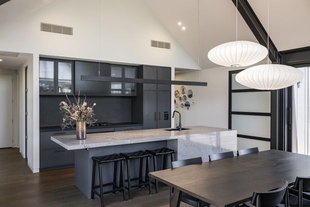 Contemporary open-plan kitchen and dining area with dark built-in cabinetry, a large pale stone island, black bar stools, and a timber dining table. The room has a high sloped ceiling, black-framed glazing and doors, and two oversized white pendant lights, creating a refined monochromatic interior with warm timber flooring.