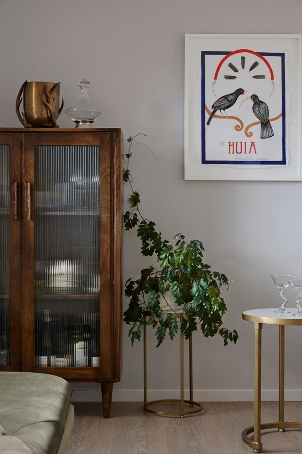 A refined living room vignette with a dark timber glass-front display cabinet, decorative ceramics and glassware, a tall indoor plant, a small brass-toned side table, and framed Huia bird artwork against a soft grey wall. Pale timber flooring and a partial upholstered sofa complete the warm, curated interior.