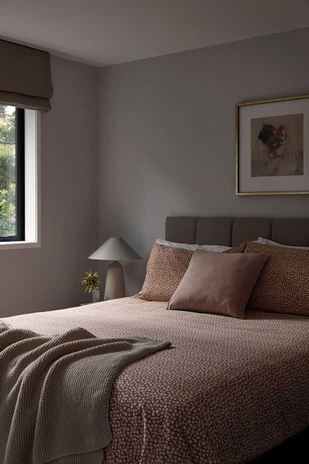 A softly lit, moody bedroom with a neatly made bed dressed in taupe and brown patterned bedding, a plush upholstered headboard, layered cushions, and a draped knitted throw. Grey-painted walls, a sculptural bedside lamp, framed artwork, and a window with a Roman blind complete the calm, refined interior.