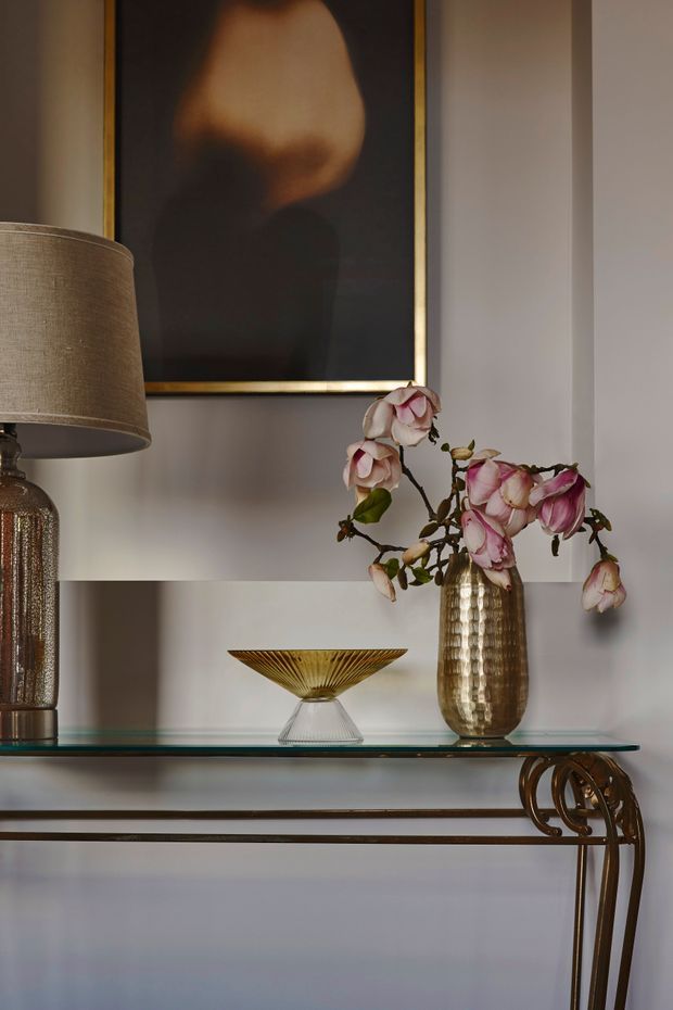 A styled interior vignette showing a glass-topped console table with ornate dark metal supports, a textured table lamp, a gold-toned vase holding pink flowers, and a decorative glass bowl. A large framed artwork with a gold frame hangs against a pale neutral wall. The composition has a refined, elegant, art-led character.