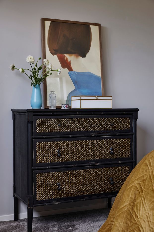 A styled bedroom vignette featuring a black three-drawer chest with ornate gold mesh drawer fronts, topped with a blue ceramic vase of white flowers, a glass vessel, and decorative boxes. A large framed abstract portrait leans against the grey wall behind the dresser, while the corner of a mustard-gold bedspread is visible at right. The scene has a refined, eclectic character with strong black, gold, blue, and mustard accents.