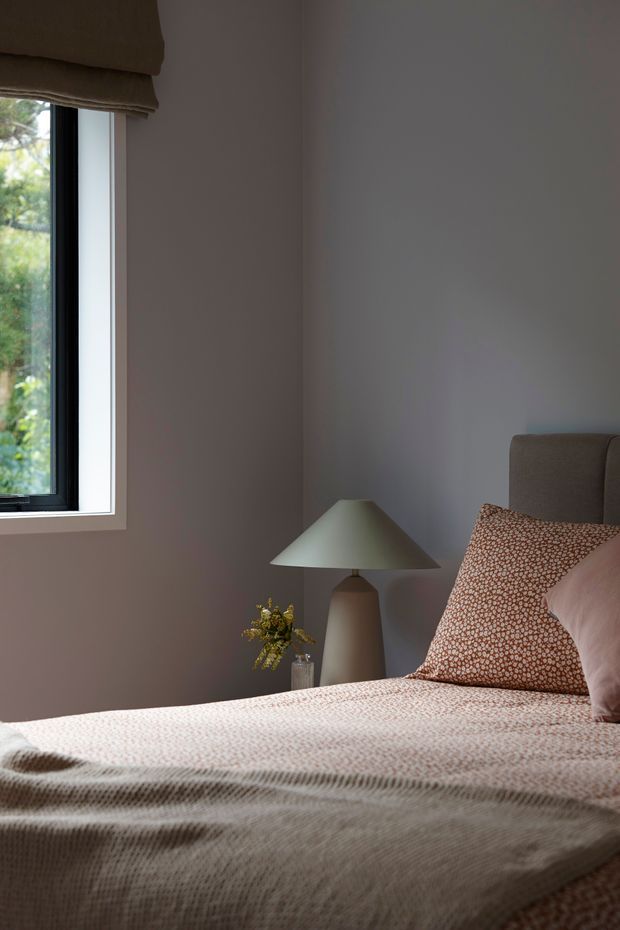 A softly lit bedroom corner with a neatly made bed, muted taupe walls, a dark-framed window with a Roman blind, and a bedside table featuring a sculptural table lamp and small vase of foliage. Patterned rust and cream bedding and cushions add warmth to the restrained, contemporary setting.
