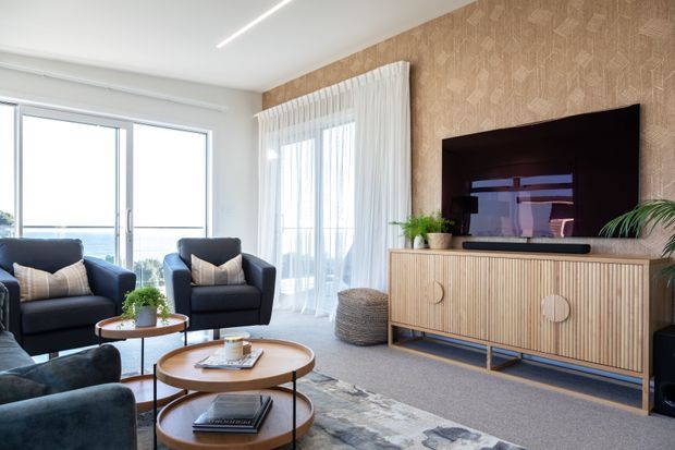 A bright, contemporary living room with large windows and sheer curtains, two dark upholstered armchairs, layered round coffee tables, and a textured area rug. A timber-toned fluted media cabinet sits against a patterned feature wall with a wall-mounted television and indoor plants. The room has a light neutral palette and a view through the glazing.