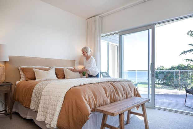 Bright, airy bedroom with a large upholstered bed dressed in warm tan and cream bedding, a simple wooden bench at the foot, and floor-to-ceiling sliding glass doors opening to a deck with an ocean view. Neutral walls, sheer curtains, and soft natural light create a relaxed coastal atmosphere.