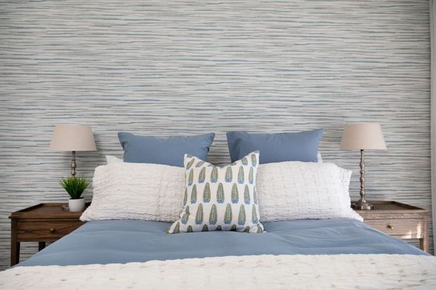 A neatly styled bedroom with a bed dressed in soft blue and white bedding, layered white and blue pillows, and a patterned accent cushion. Matching bedside tables with lamps flank the bed, while the wall behind features textured horizontal striped wallpaper.
