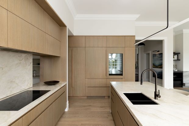 A refined contemporary kitchen with full-height light timber cabinetry, a pale natural-stone splashback and island benchtop, integrated black cooktop and twin sinks, and a slim black pendant light. The kitchen opens toward an adjoining living area, with detailed white ceiling cornices and a warm, restrained material palette.