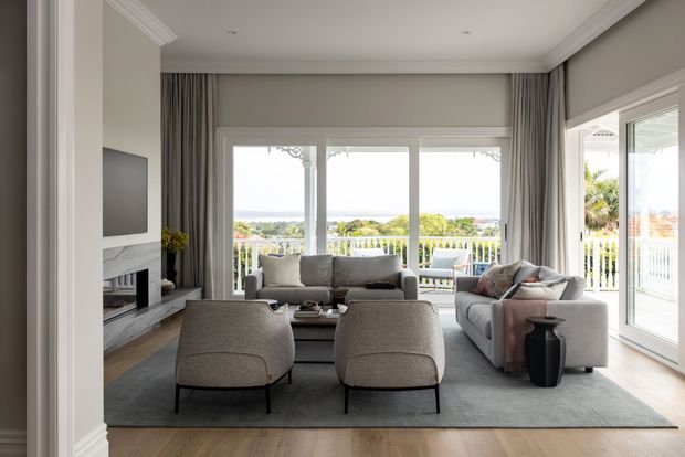 A bright, refined living room with a neutral grey palette, a large sectional sofa, two upholstered lounge chairs, a dark coffee table, and a built-in fireplace and television wall. Floor-to-ceiling glazing and wide balcony doors open to a white balustrade and expansive distant views. Full-height curtains, recessed ceiling lights, decorative cornices, and timber flooring contribute to a polished contemporary interior.