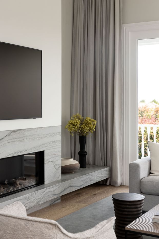 Refined contemporary living room with a marble-clad fireplace and integrated hearth ledge beneath a wall-mounted television. Full-height grey curtains frame a bright glazed door or window, while neutral upholstered seating, a dark sculptural side table, and understated decorative objects create a calm, luxurious scheme. Timber flooring and a soft grey rug add warmth and texture.