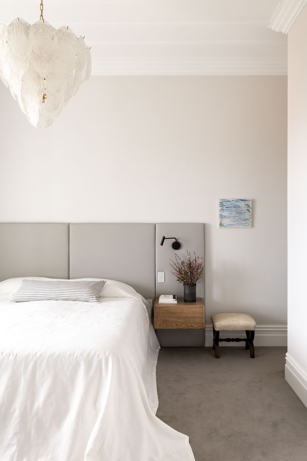 A refined bedroom with a large upholstered grey bedhead, white bedding, grey carpet, warm neutral walls, and crisp white decorative ceiling cornices and skirting. A timber bedside drawer with integrated wall lamp sits beside the bed, complemented by a small upholstered stool, floral arrangement, artwork, and an elegant sculptural pendant light. The overall design is calm, understated, and softly luxurious.