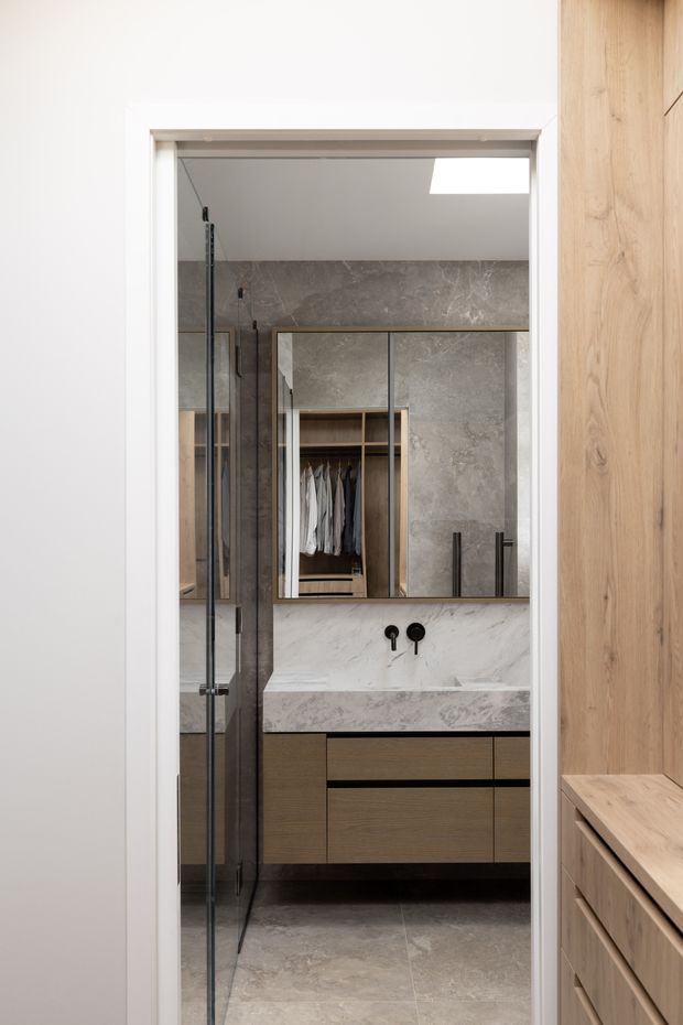 A refined bathroom viewed through an open doorway, featuring a glass shower enclosure, large grey stone-look wall and floor tiles, a white marble vanity splashback and benchtop, dark timber floating cabinetry, black tapware, and mirrored storage above the vanity. A timber wardrobe or cabinetry zone is visible at the right edge and reflected in the mirror.