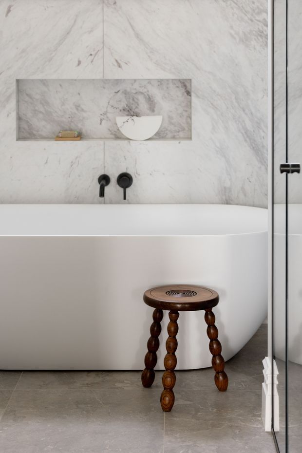 A refined contemporary bathroom featuring a freestanding white bathtub set against full-height veined marble wall panels. A recessed marble shelf holds a sculptural bowl, with matte black wall-mounted tapware and a glass shower partition at right. The grey stone-look floor and dark carved timber stool add contrast and warmth.