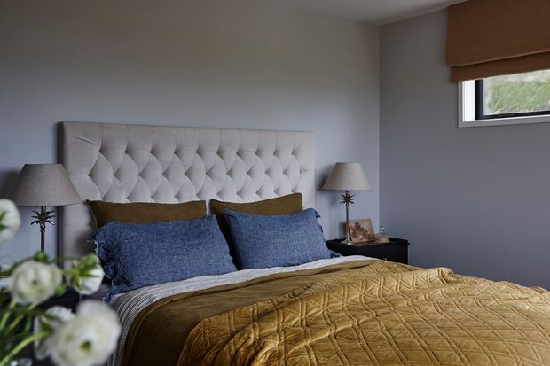 A cozy bedroom with a large tufted upholstered headboard, layered blue and ochre bedding, matching bedside tables and lamps, and a small high-set window with a brown roller blind. The room has muted grey walls and a warm, restful atmosphere.