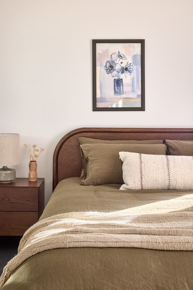 A warmly styled bedroom featuring a dark wood bed with a rounded headboard, olive-brown linen bedding, a textured cream throw, and a matching dark wood bedside table. A ceramic lamp and small vase sit beside the bed, while framed floral artwork hangs on the neutral wall. The room has a calm, earthy, contemporary-traditional feel with soft natural light.