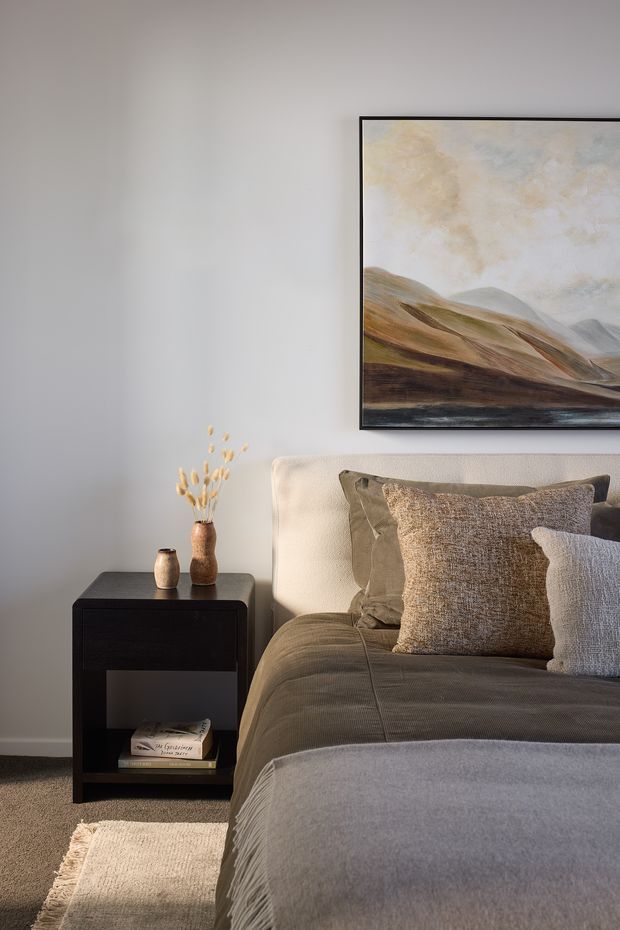 A softly lit, contemporary bedroom corner with a neatly made bed, layered neutral bedding and textured cushions, a dark bedside table with ceramic vessels and dried stems, and a large framed landscape artwork above the bed. The palette is warm and muted, with pale grey walls, taupe textiles, dark brown carpet, and a cream rug.