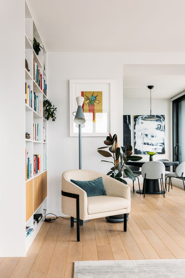 Bright, refined open-plan living and dining area with white walls and built-in shelving filled with books and decorative objects. A textured cream lounge chair with a dark teal cushion sits on pale timber flooring beside a sculptural floor lamp and indoor plants. The background contains a round dining table with upholstered chairs, a pendant light, large artwork, and black-framed glazing. The space has a clean contemporary feel with mid-century-inspired furniture accents.