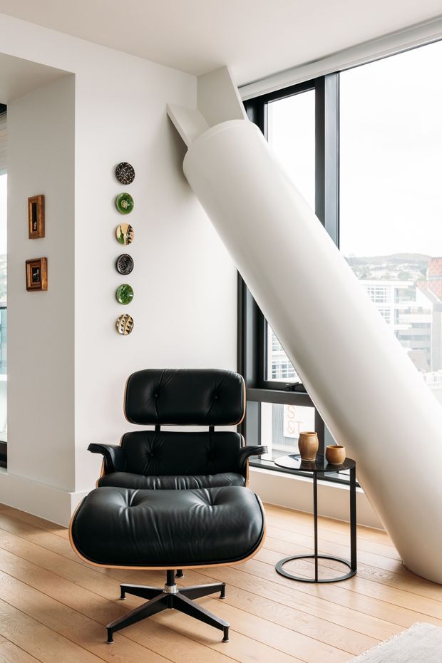 Bright contemporary interior corner with a black leather lounge chair and matching ottoman, positioned beside a slim black side table and floor-to-ceiling windows. A large sculptural white diagonal architectural element dominates the space, with light timber flooring, white walls, framed artwork, and decorative wall plates. Urban buildings are visible outside the glazing.