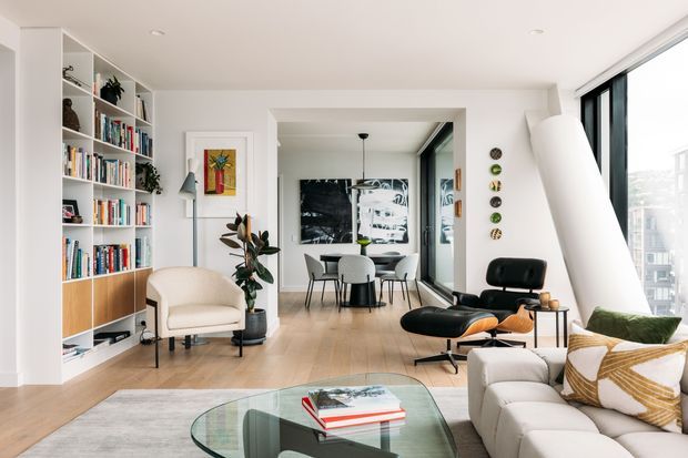 Bright, open-plan contemporary living room with floor-to-ceiling glazing, pale timber flooring, extensive built-in shelving, a cream sofa and armchair, and a sculptural white angled feature wall or architectural element. A dining area is visible beyond the living space, with modern furniture, indoor plants, artwork, and a glass coffee table.