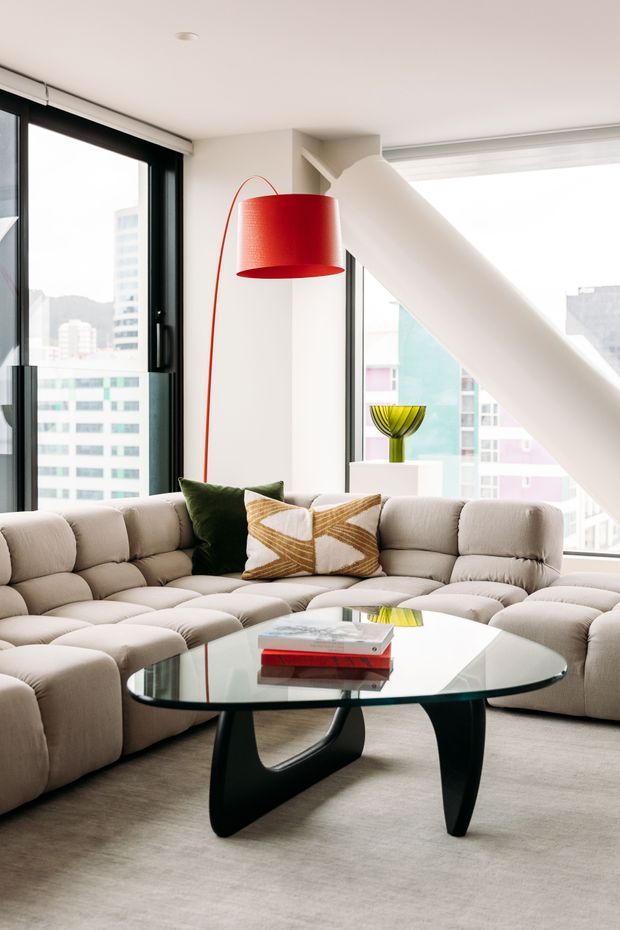 Bright contemporary living room in a high-rise apartment, featuring floor-to-ceiling black-framed windows, a large curved modular sectional sofa, sculptural black glass coffee table, red pendant lamp, and a prominent white diagonal structural beam. City buildings are visible through the glazing.