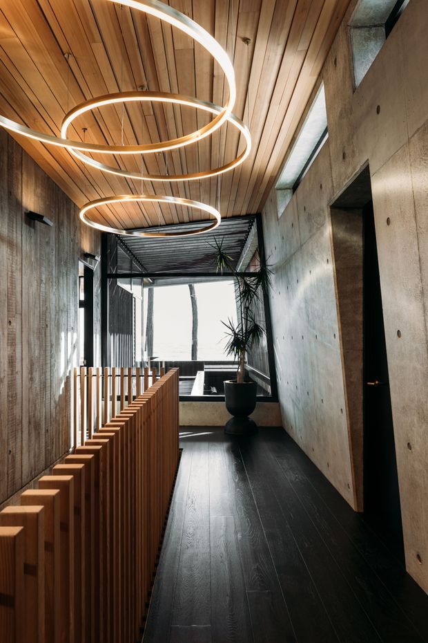 A narrow, dramatic interior corridor or landing with a dark timber floor, exposed concrete walls, and a warm timber slatted ceiling. Sculptural circular pendant lights run overhead, while a timber balustrade lines the open edge. Full-height glazing and a planted courtyard are visible at the far end, creating a strong indoor-outdoor connection. The palette is predominantly warm wood, charcoal, black, and raw grey concrete.
