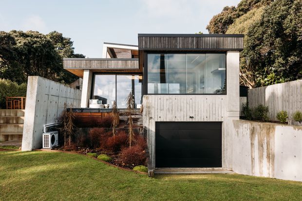 Exterior view of a contemporary multi-level home set into a landscaped sloping site. The facade combines light vertical timber cladding, large glazed openings, concrete retaining walls, projecting balconies, and an integrated dark garage door. Mature trees and lawn surround the building.