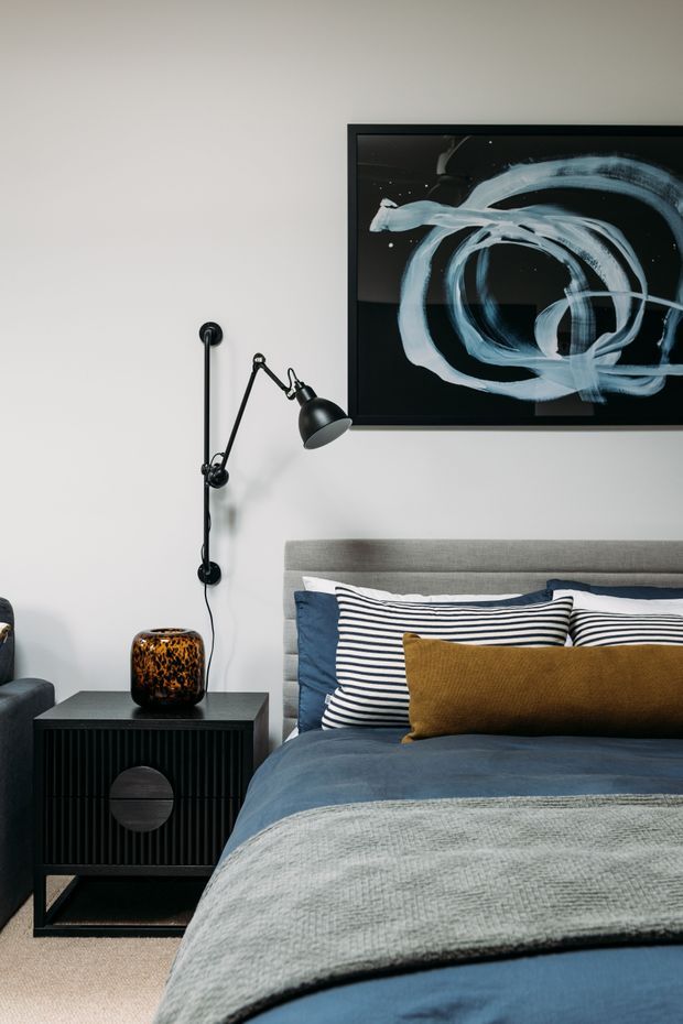 A styled bedroom featuring a grey upholstered bedhead, layered blue, grey, mustard and striped bedding, a black fluted bedside table, and a black articulated wall-mounted reading lamp. A large abstract black-and-white artwork hangs above the bed against a light neutral wall.