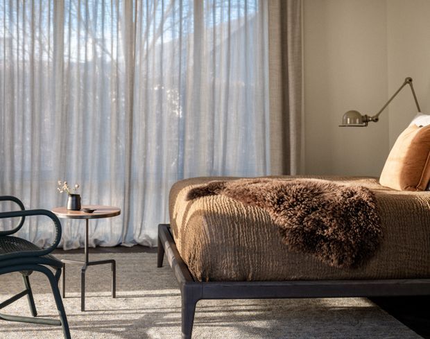A warmly lit, contemporary bedroom with a low dark timber bed, textured brown bedding and a dark brown faux-fur throw. Floor-to-ceiling sheer curtains filter natural light across a neutral wall and carpeted floor. A sculptural black lounge chair, small round side table, and articulated wall-mounted reading lamp complete the restrained, cozy interior.