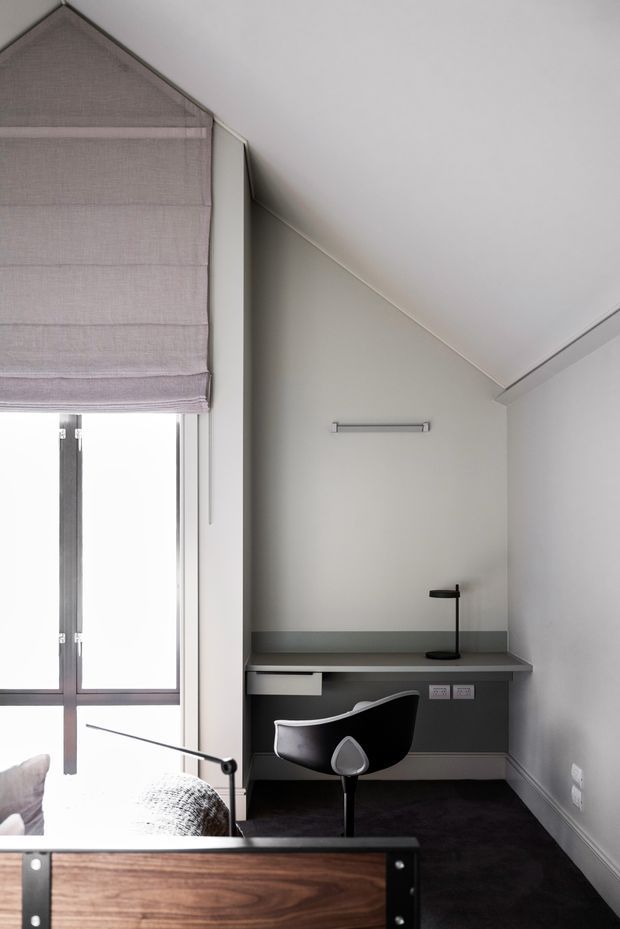Compact contemporary study nook in a sloped-ceiling room, featuring a built-in dark desk with integrated drawers, a black task chair, slim wall light, dark carpet, pale grey walls, and a large window with a soft mauve Roman blind. Part of a bed and a wooden chest are visible in the foreground, indicating the workspace is within a bedroom.