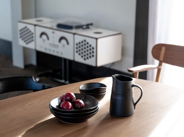 A warm, softly lit dining area with a light timber dining table, black ceramic bowls, a black pitcher, and red fruit in the foreground. A wooden armchair and a distinctive retro-style white and black audio console are visible against a pale wall in the background.