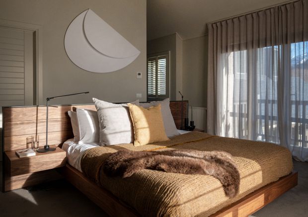 Warm, refined bedroom with a low timber bed and integrated wood headboard, layered neutral bedding, bedside task lighting, sculptural wall art, plantation shutters, and full-height glazed doors dressed with sheer curtains. Strong afternoon sunlight creates a cozy, sophisticated atmosphere.