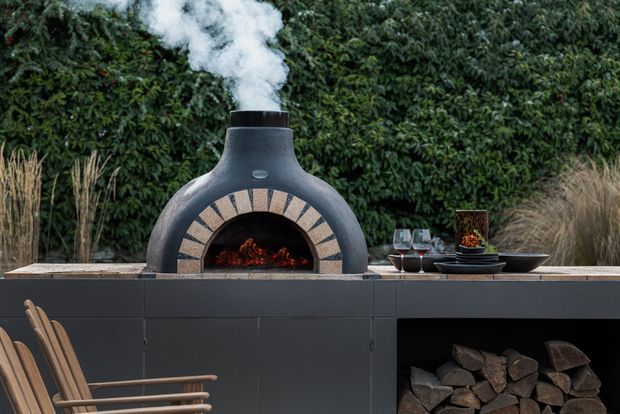 A stylish outdoor entertaining area featuring a freestanding dark charcoal wood-fired pizza oven with smoke rising from its chimney, set into a dark built-in outdoor kitchen counter. Firewood is stored beneath the counter, with wine glasses and serving ware arranged beside the oven. A dense green hedge forms the backdrop, and timber outdoor chairs are partially visible in the foreground.