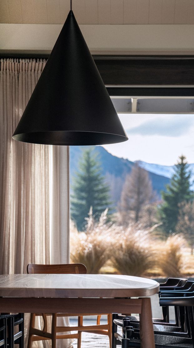 A warm, refined dining area with a large black cone pendant light suspended above a timber dining table. Floor-to-ceiling curtains frame expansive glazing with mountain and wooded scenery beyond, while dark dining chairs and a dark window surround add contrast. The room has a mid-century-influenced, natural material palette and strong indoor-outdoor connection.