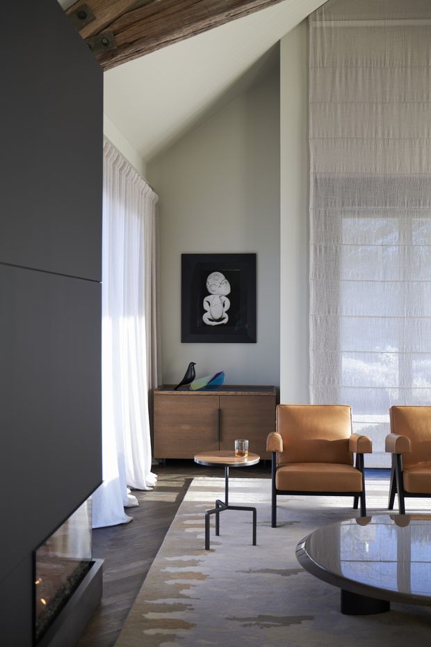 A refined living room with a dramatic vaulted ceiling, exposed rustic timber beam, tall sheer window treatments, and a dark built-in fireplace wall at left. The room features a mid-century-inspired timber sideboard, leather lounge chairs, nesting side table, round coffee table, neutral textured rug, and understated decorative objects. Natural light creates a calm, warm and sophisticated atmosphere.