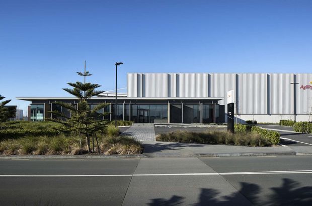 Exterior view of a contemporary commercial or industrial building with a large corrugated metal-clad volume, dark glazed entry frontage, a low flat-roofed wing, landscaped forecourt, paved pedestrian entrance, and adjacent roadway under clear daylight.