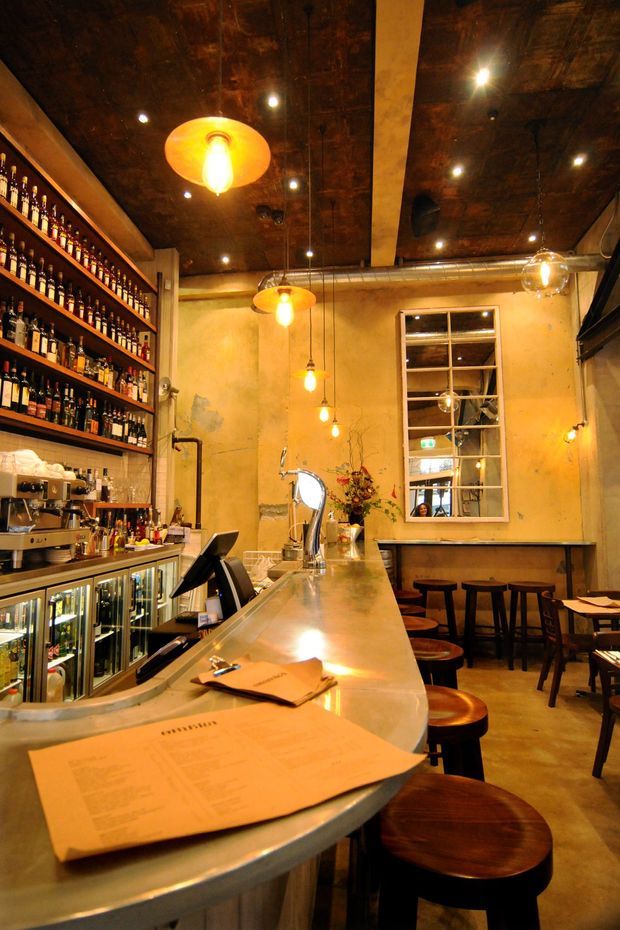 Warm, atmospheric interior of a compact industrial-style bar or restaurant. A long curved polished counter with stools dominates the foreground, backed by illuminated glass-front beverage fridges, an espresso machine, and extensive open shelving filled with bottles. The space features aged textured plaster walls, a dark timber ceiling with exposed services, suspended pendant lights, a large industrial-framed mirror, and a small rear dining area with bar-height tables and stools.