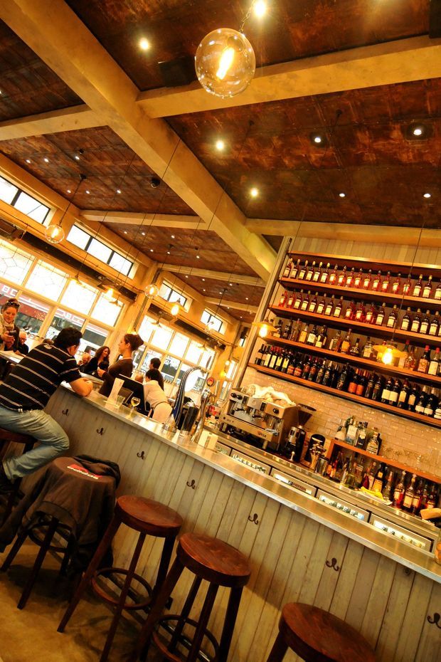 A warmly lit commercial bar interior viewed at an angle, with a long timber-clad counter and wooden bar stools in the foreground. Behind the counter are a coffee machine, taps, glassware, and extensive open shelving displaying bottles. The space features exposed timber ceiling panels, large pale structural beams, recessed spotlights, pendant globe lights, tiled backsplash, and generous street-facing windows. Patrons are seated at the bar, reinforcing the hospitality setting.