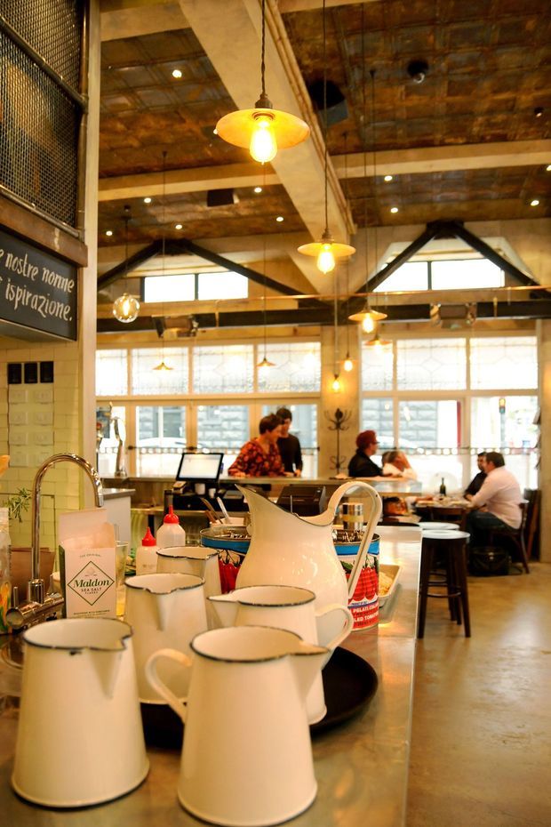 Warmly lit industrial-style café interior viewed from behind a service counter. The space features exposed timber and steel roof structure, concrete columns, large gridded windows, hanging pendant lights, dark metal framing, and patrons seated at tables in the background. White enamel pitchers and café supplies occupy the foreground counter.