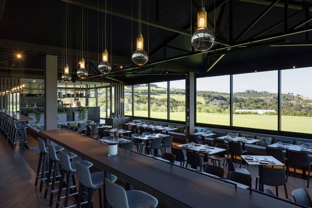 A spacious contemporary restaurant or hospitality dining interior with extensive floor-to-ceiling glazing overlooking a green rural landscape. The room features long communal counters with high stools, multiple neatly arranged dining tables, dark finishes, exposed black structural framing and services, and a row of distinctive suspended globe-and-lantern pendant lights.