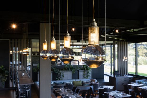 A dark, refined contemporary restaurant or hospitality interior with dramatic hanging pendant lights, floor-to-ceiling glazing, exposed dark ceiling elements, and neatly arranged dining tables. A bar or service counter, lounge seating, indoor planting, and views to a landscaped exterior are visible.