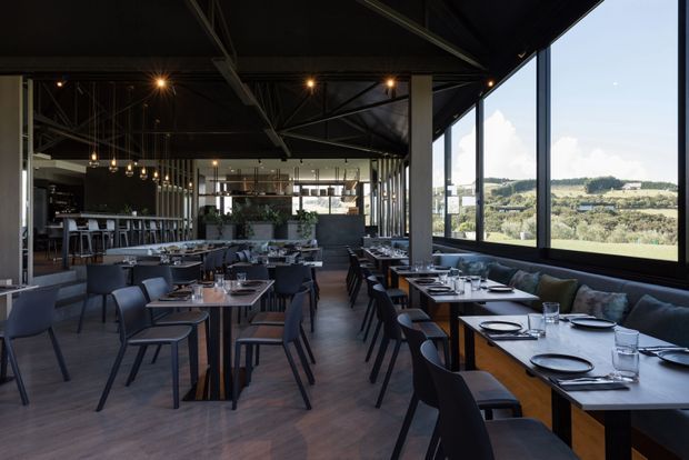 A spacious contemporary restaurant interior with rows of dark timber dining tables and black upholstered chairs, a long banquette along full-height windows, a visible bar and open dining layout, and an exposed dark structural ceiling with track and pendant lighting. Large glazing overlooks a green rural or suburban landscape.