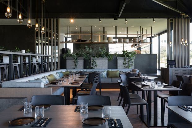 A sophisticated contemporary restaurant interior with a central lounge seating area, multiple dark timber dining tables, upholstered banquette seating, a bar to the left, and a large open kitchen or service counter at the rear. Black structural elements, suspended pendant lights, metal-framed partitions, concrete planter boxes, and abundant greenery create an industrial yet refined atmosphere.