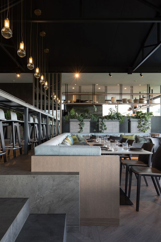 A sophisticated contemporary restaurant interior with a dark industrial-style ceiling, exposed structural framing, black metal screens and railings, and dramatic globe pendant lights. The dining area features upholstered banquette seating, timber dining tables, dark chairs, concrete-clad stepped platforms, and large built-in planters filled with trailing greenery. A raised bar or mezzanine-like area is visible in the background.