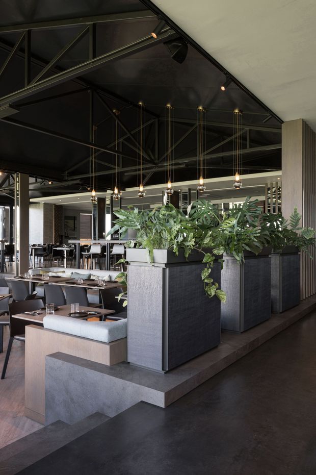 A sophisticated contemporary restaurant interior with dark industrial-style ceiling structure, exposed black beams, suspended pendant lights, and extensive integrated planter boxes dividing the dining areas. The space features dark polished flooring, timber-toned dining tables, upholstered banquette seating, black dining chairs, and textured charcoal planter cabinetry. Vertical timber slats and reflective surfaces add depth to the refined hospitality fit-out.