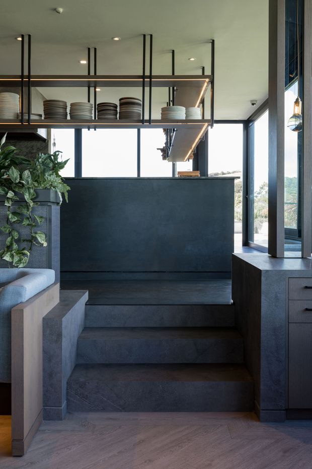 A refined, dark-toned interior showing a raised kitchen or dining counter area reached by broad concrete-clad steps. Black-framed glazing, a deep charcoal island or counter, open metal shelving with stacked crockery, timber cabinetry, integrated warm lighting, and a leafy indoor plant create a contemporary restaurant-like kitchen setting. The space opens toward an exterior view through large windows.