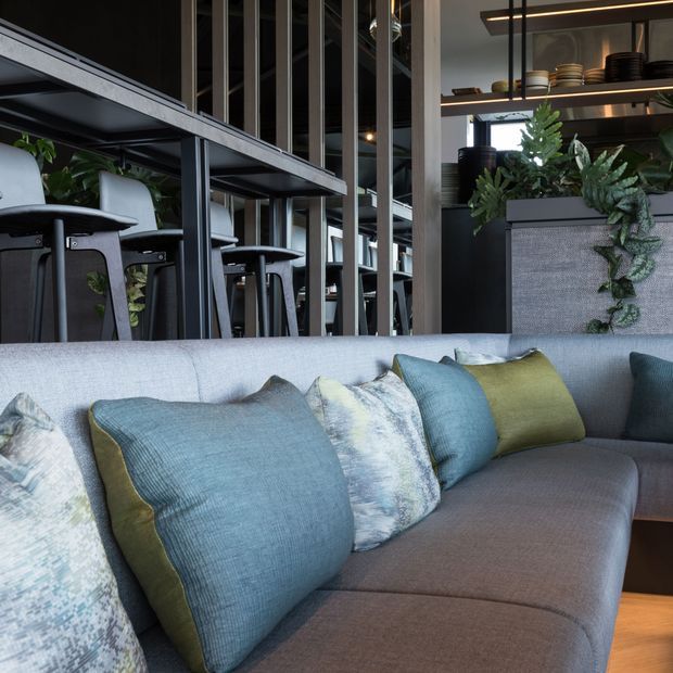 Contemporary commercial hospitality interior featuring a grey upholstered sectional sofa with blue, olive, and patterned cushions in the foreground. Behind it is a dark bar or dining area with black metal-framed shelving, high stools, stacked tableware, integrated warm lighting, vertical metal screens, and cascading greenery.