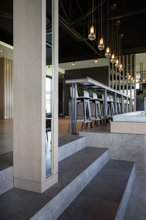 A dramatic contemporary interior with a dark exposed structural ceiling, black walls, and a long high communal table with bar stools. A line of suspended globe pendant lights hangs above the table, while broad concrete-clad steps create tiered seating in the foreground. Vertical timber-toned wall panelling and slender dark metal framing add architectural definition.