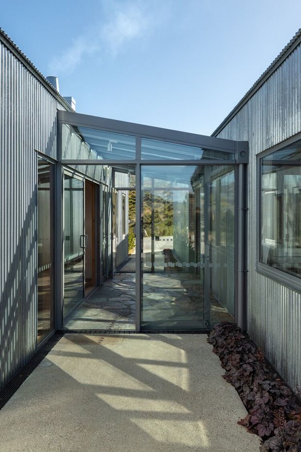 A narrow glazed passageway between two corrugated metal-clad wings of a contemporary building. Dark aluminium-framed glass doors and a glazed roof connection create a bright enclosed threshold, with exposed stone paving visible beyond and linear planting along the concrete walkway. The composition emphasizes the modern industrial material palette, strong geometric lines, and indoor-outdoor connection.