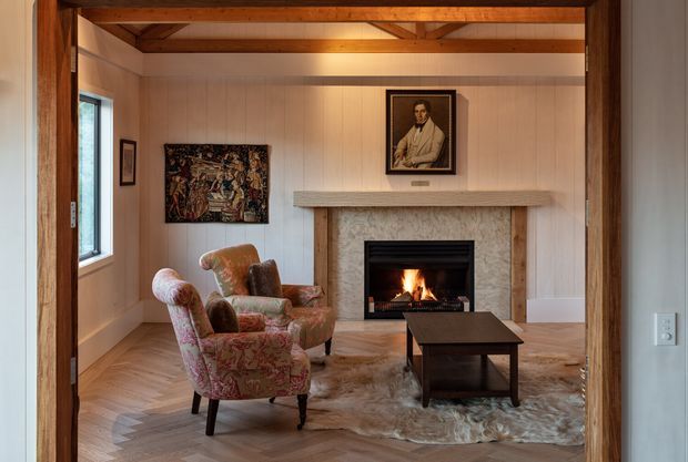A warm, traditional living room viewed through a timber-framed opening. The room features pale vertical wall panelling, exposed timber ceiling beams, a stone fireplace with a lit fire and mantel, vintage upholstered armchairs, a dark coffee table, herringbone timber flooring, and a hide rug. Artwork and a framed portrait add a refined heritage character.