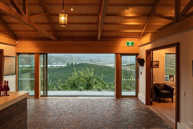Warm timber-lined interior with exposed post-and-beam ceiling structure, extensive timber-framed glazing overlooking a vineyard and mountainous landscape, and a dark brick-paver floor. A built-in timber counter with wine bottles occupies the left foreground, while an open doorway on the right leads to a small furnished sitting area. A pendant lantern and emergency exit sign are visible.
